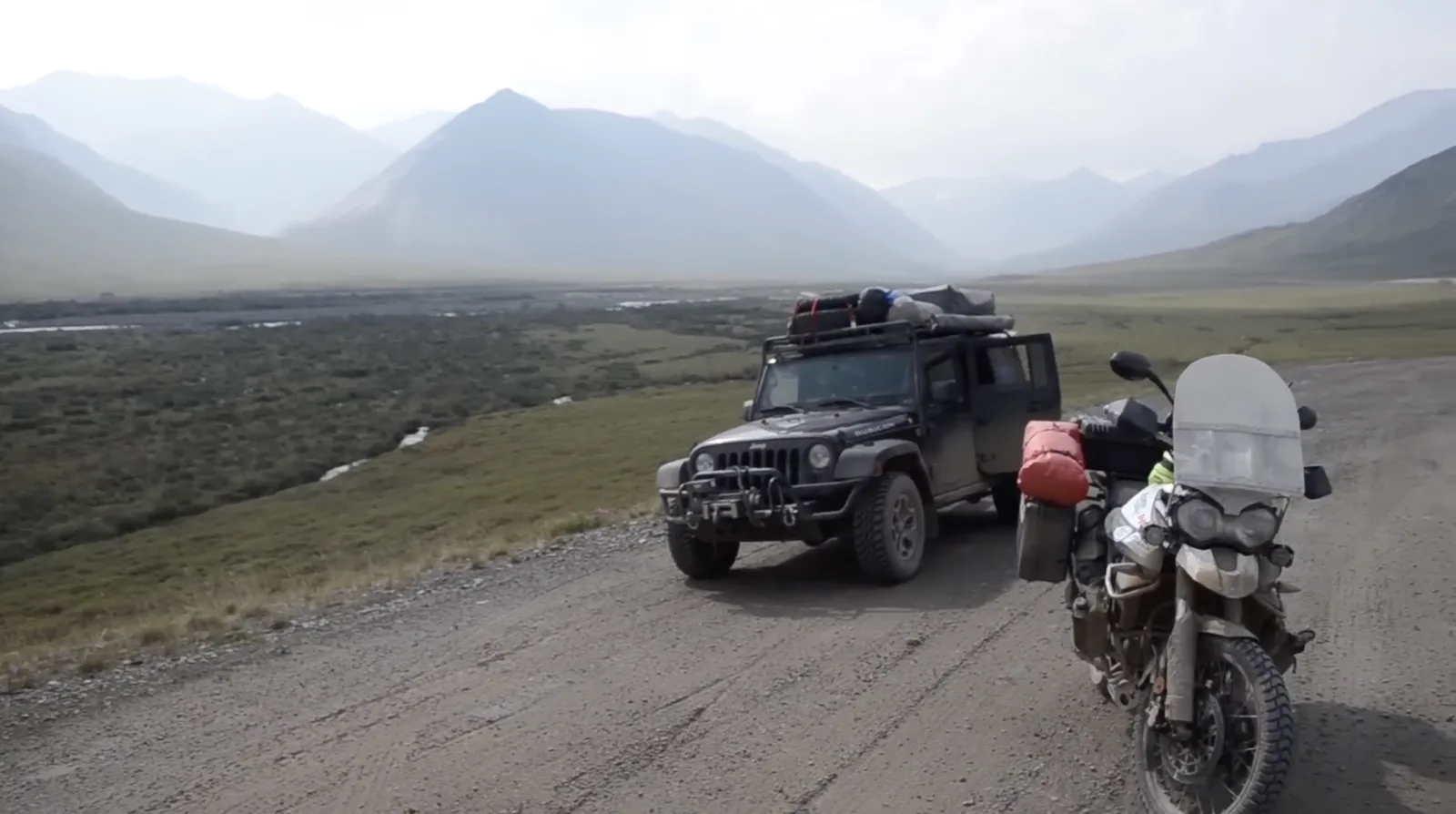 A loaded Jeep and an adventure motorbike parked on a gravel road in a wide, empty mountain valley.