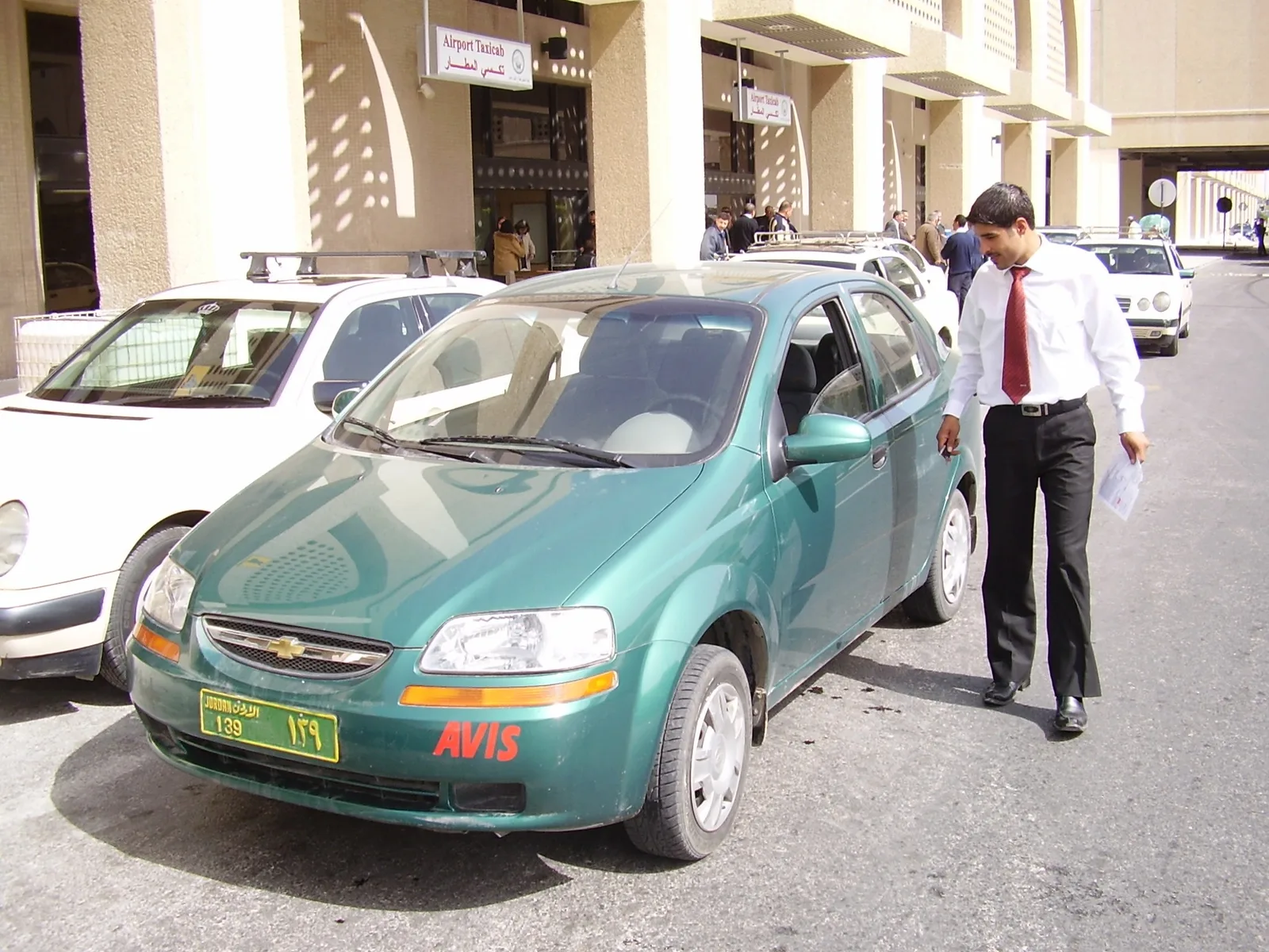A green Avis rental car outside an airport terminal, with a man in a shirt and tie walking beside it.