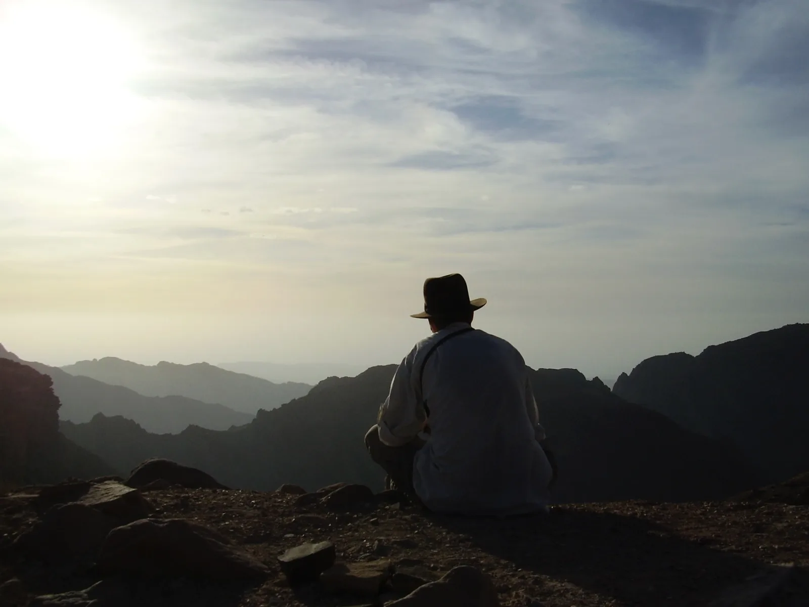 A person in a wide-brimmed hat sitting alone on a summit at sunrise, looking out over layered mountain ridges.