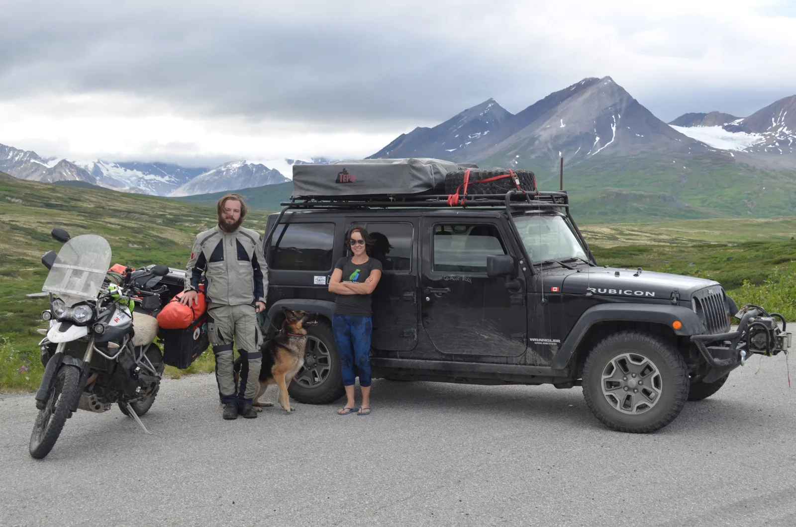 A man, woman, and dog standing in front of a mountainscape with an expedition Jeep and Motorcycle.
