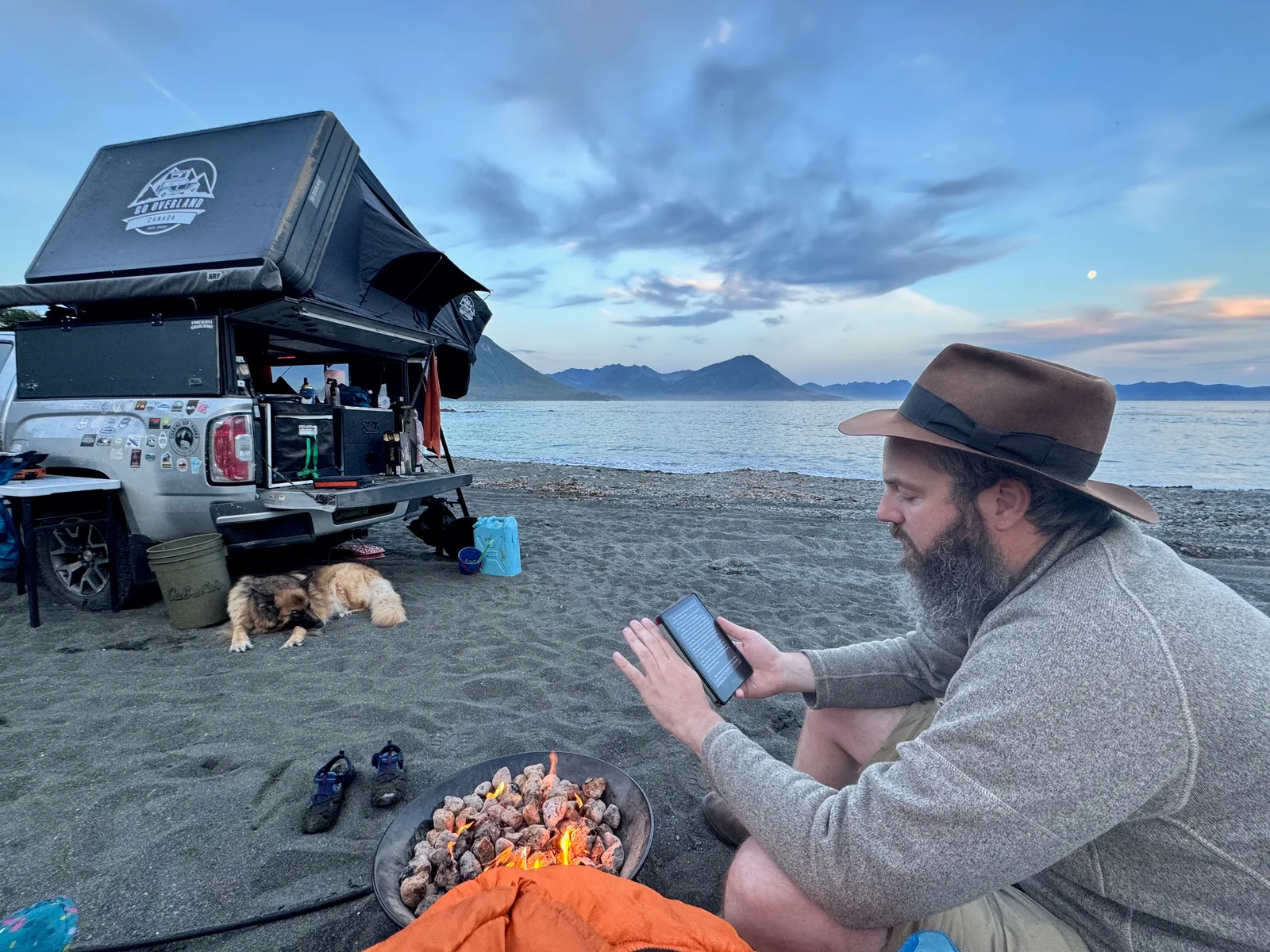 A truck with a roof tent, a dog sleeping under the tailgate, and a man in a hat reading an e-reader by a fire with an ocean and mountains in the background.