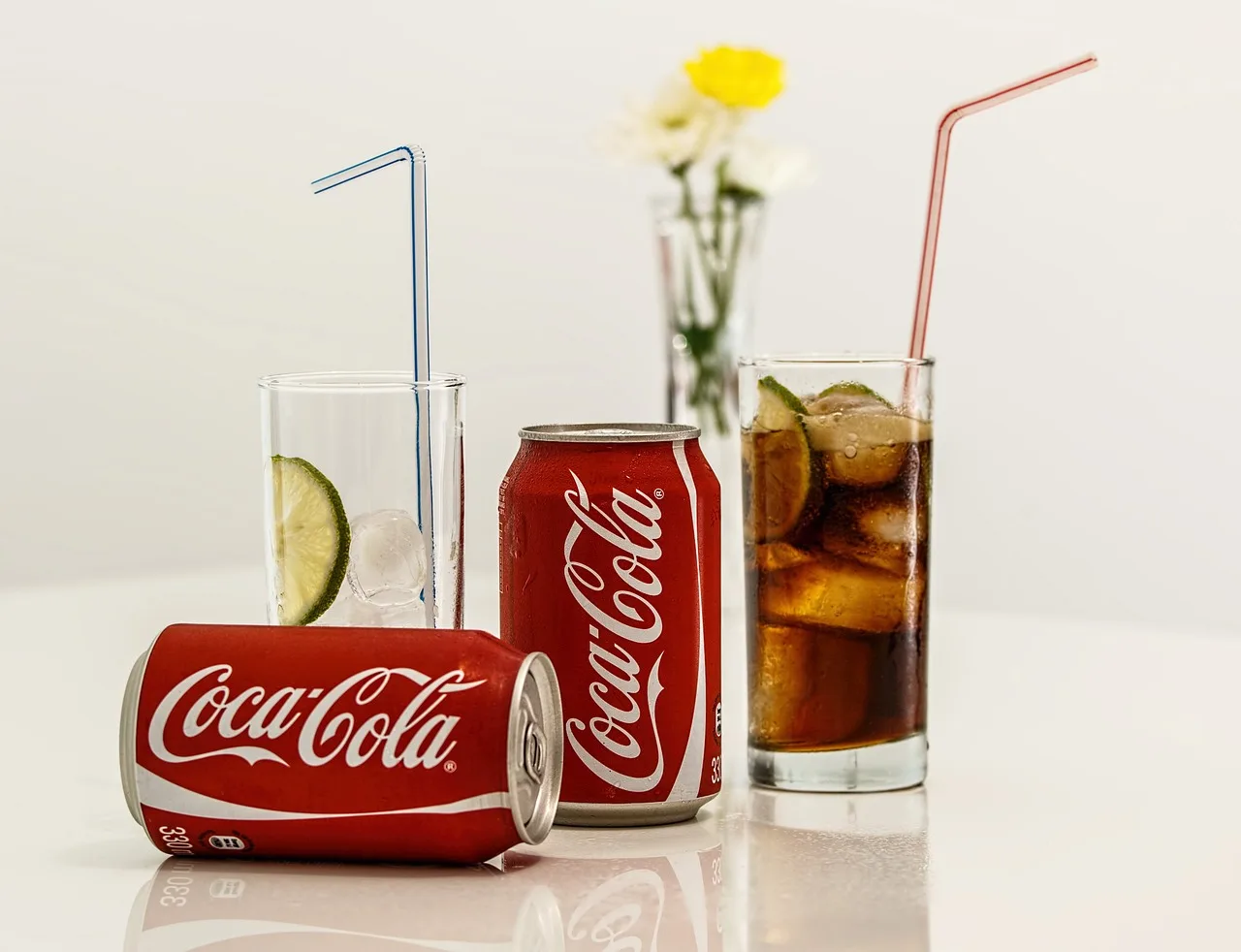 Two cans of Coca-Cola beside a tall glass of cola with ice and a slice of lime.