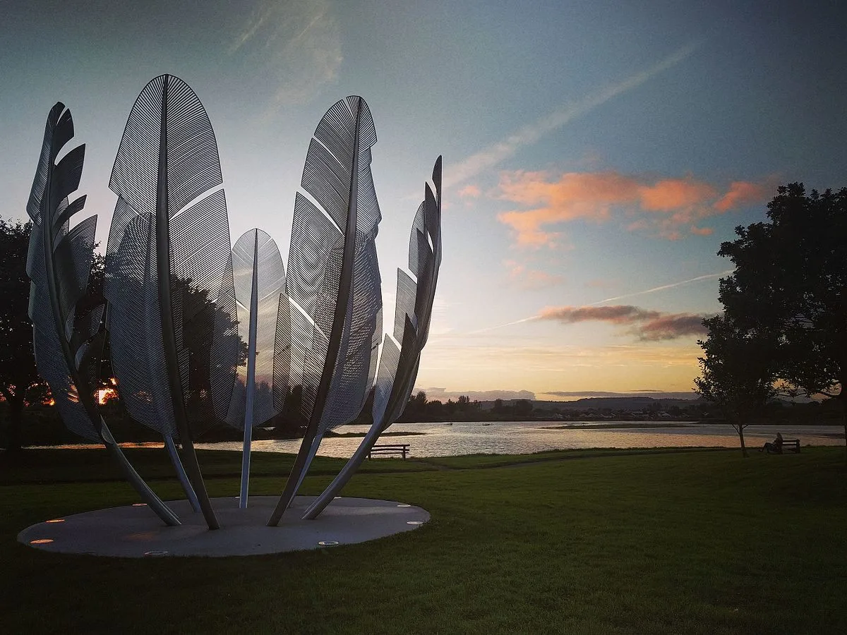 A statue of massive feathers, pointing upwards to the sky and arranged in a circle with the sun rising in the background.