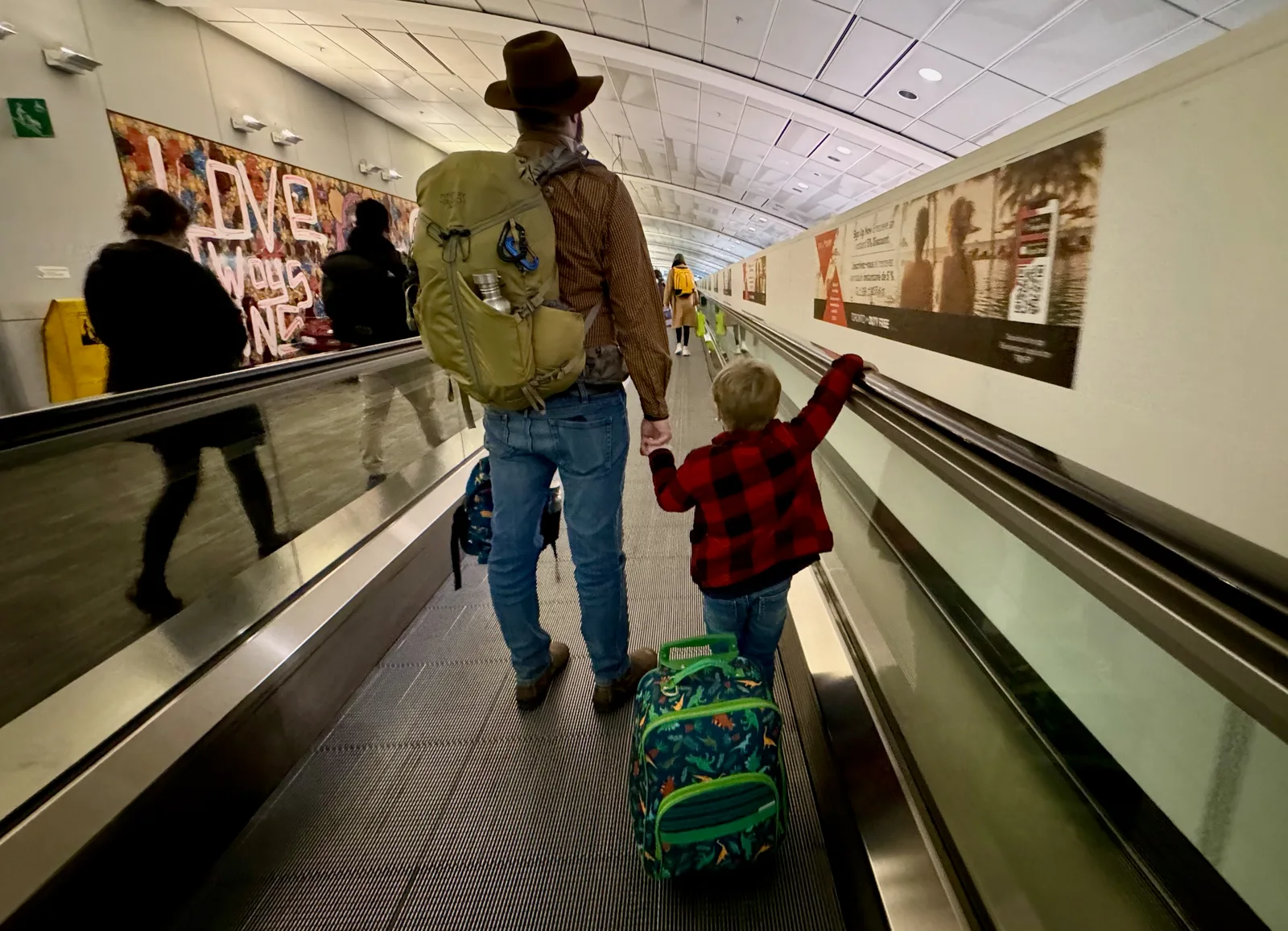 A father wearing a backpack and holding his son's hand on a moving sidewalk at the airport.