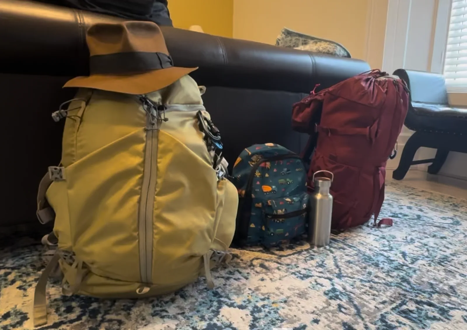 Two adult backpacks and a child'ss backpack at the end of a bed.
