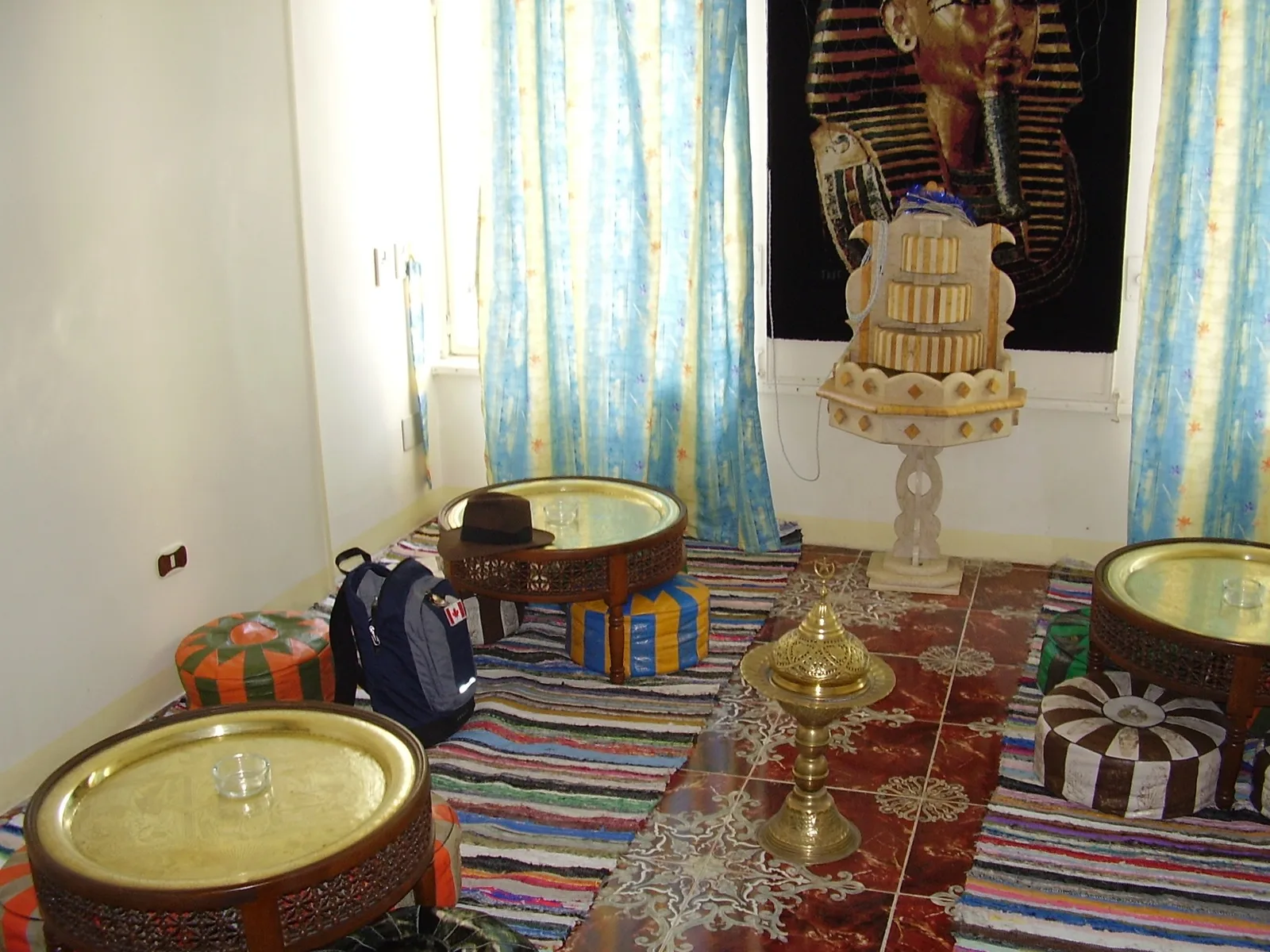 An Egyptian tea room with small cushions for sitting, a backpack leaning against one cushion, and a picture of Tut's sarcophagus on the wall with a vibrant carpet on the floor.