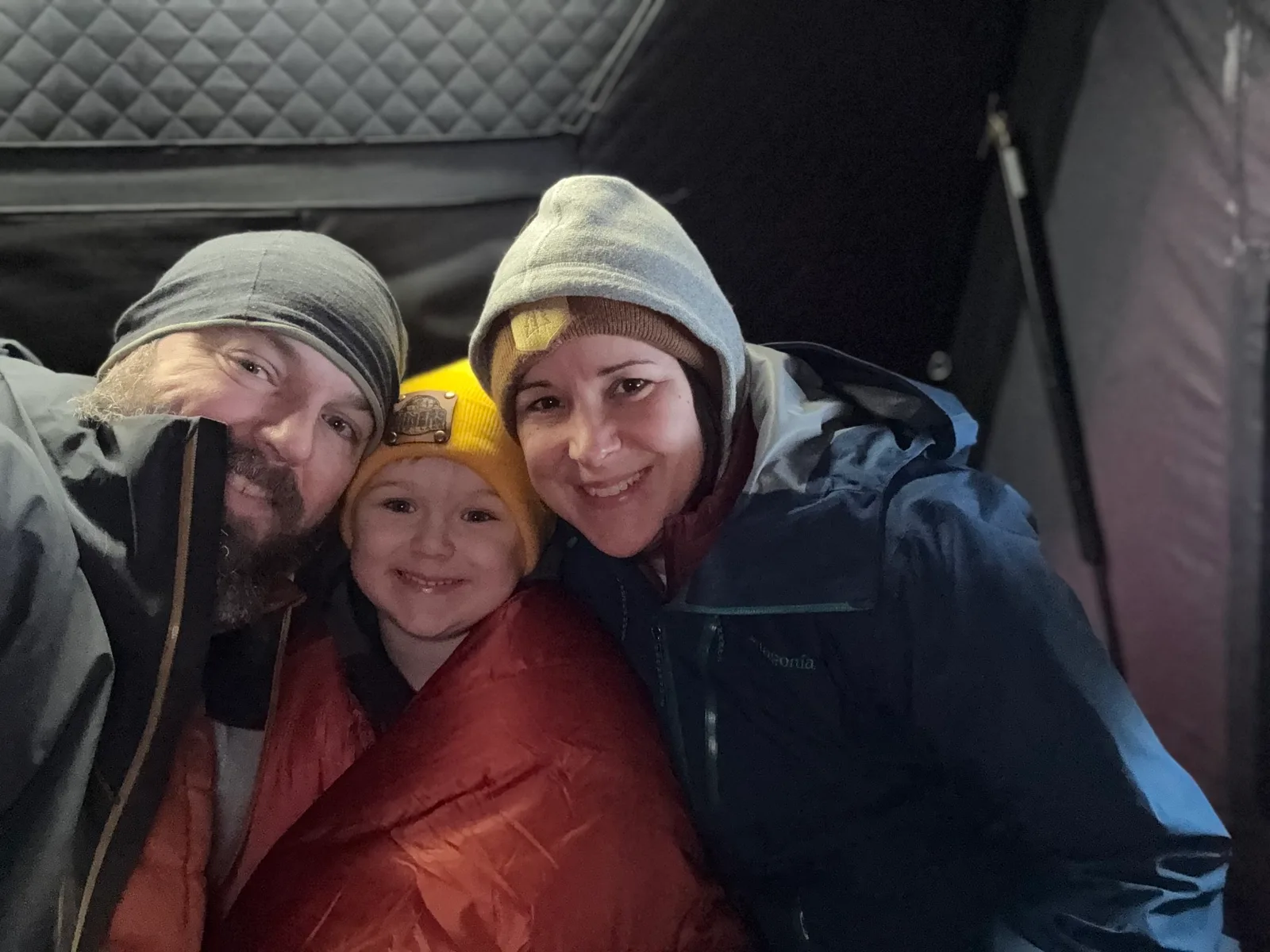 A man, a woman, and a child bundled in sleeping bags and winter gear, apparently inside a tent. They appear to be camping, but they are smiling broadly at the camera - clearly enjoying their time!
