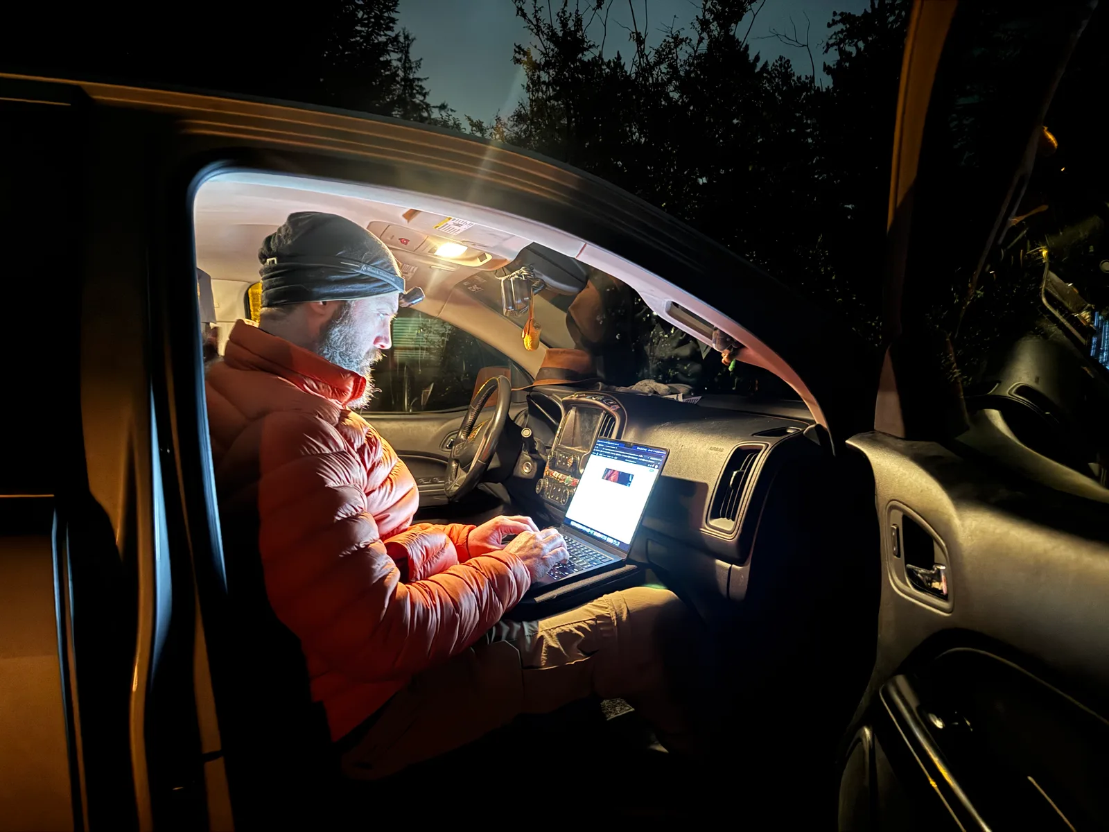 A man sitting in the passenger seat of a truck wearing a toque, and orange puffy coat, and a headlamp while writing on his laptop.
