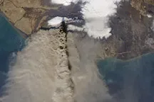 A satellite view of a volcanic ash plume drifting from a coastline out over the sea.