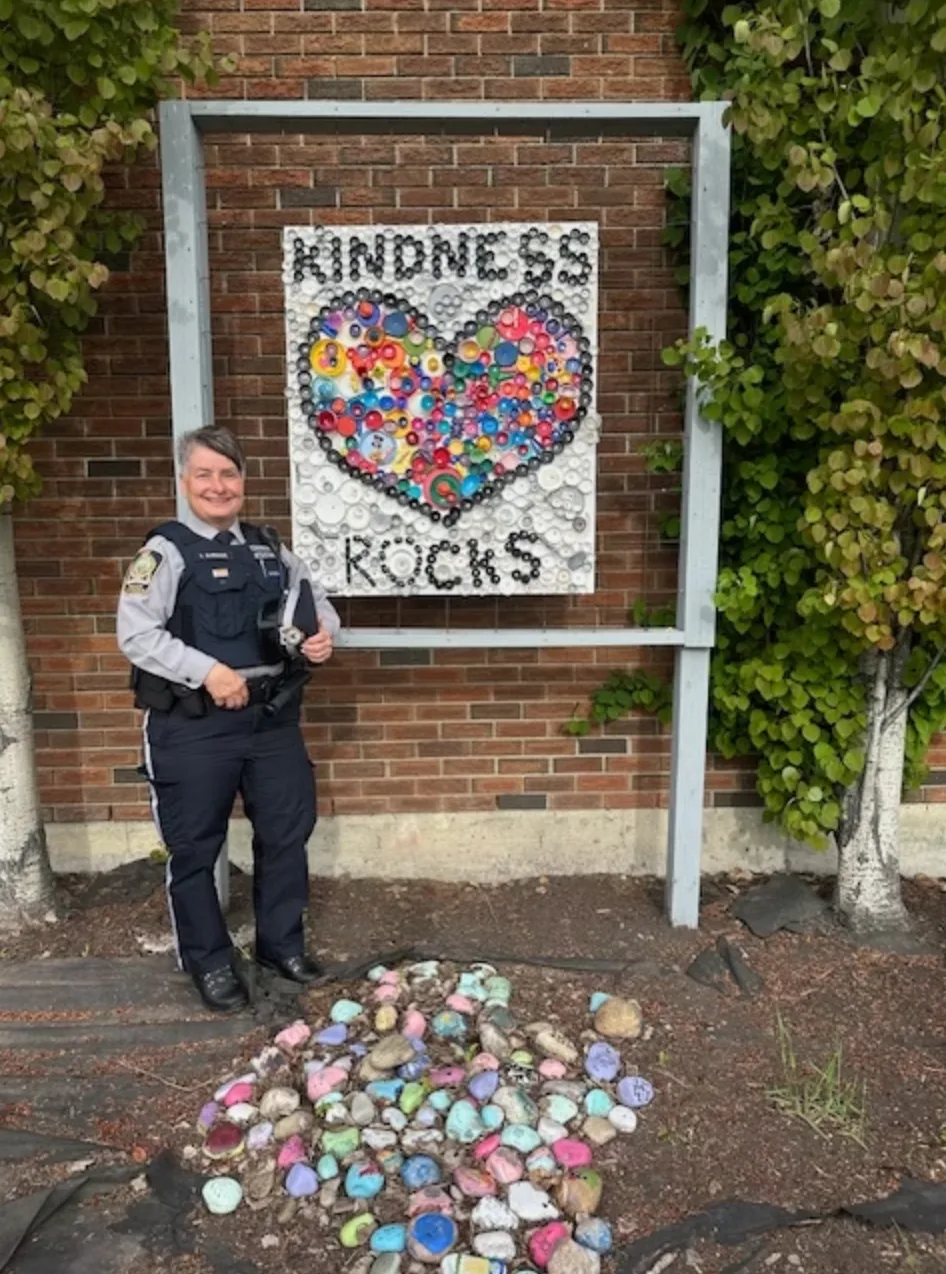 A uniformed police officer standing beside a "Kindness Rocks" sign, with a bed of brightly painted stones laid out at its base.