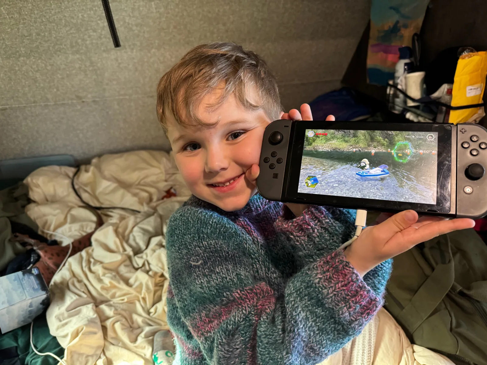 A young child sitting on a bed inside a camper, smiling and holding up a Nintendo Switch.