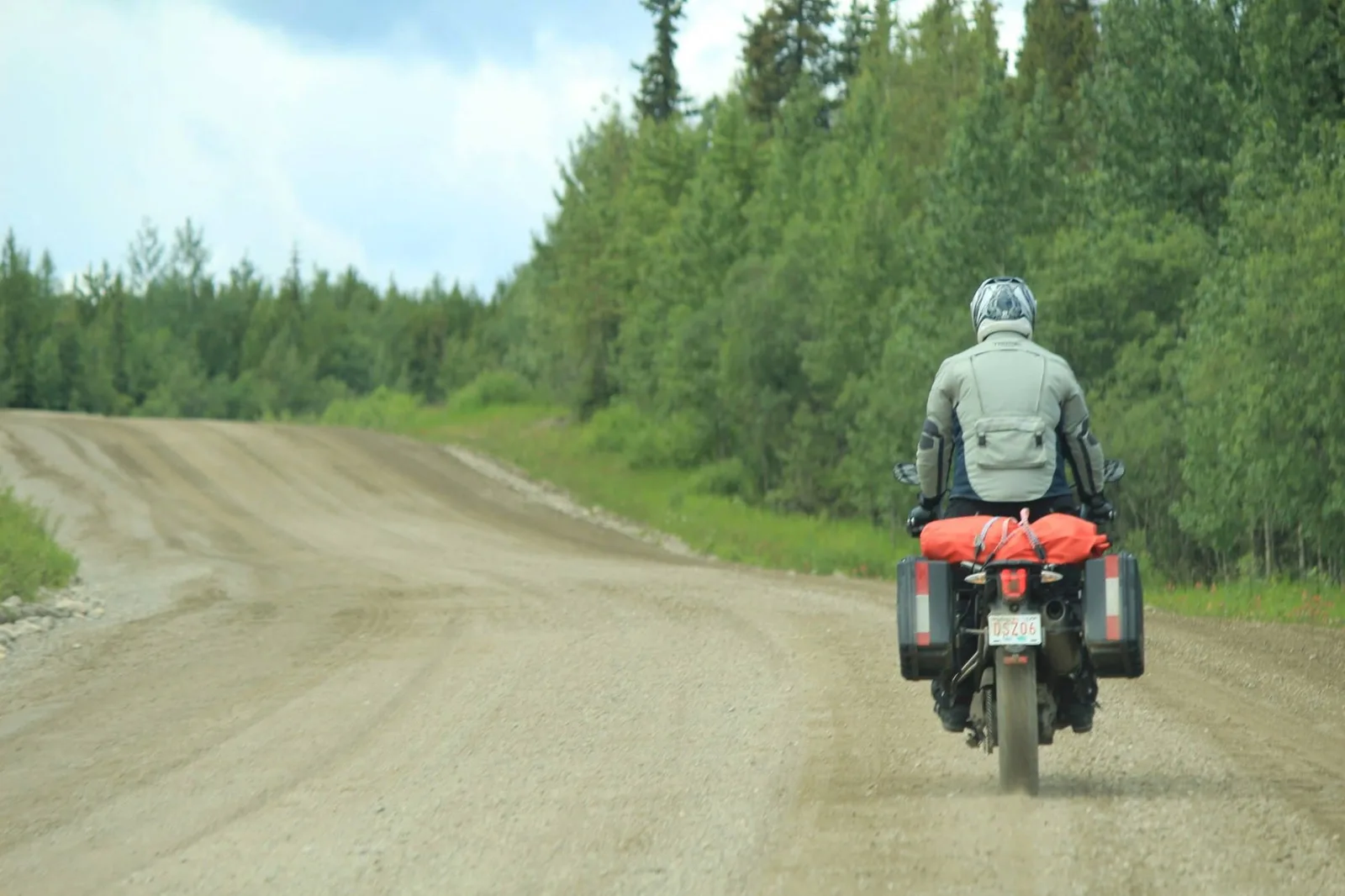 A motorcyclist riding away along a gravel road cut through thick boreal forest.