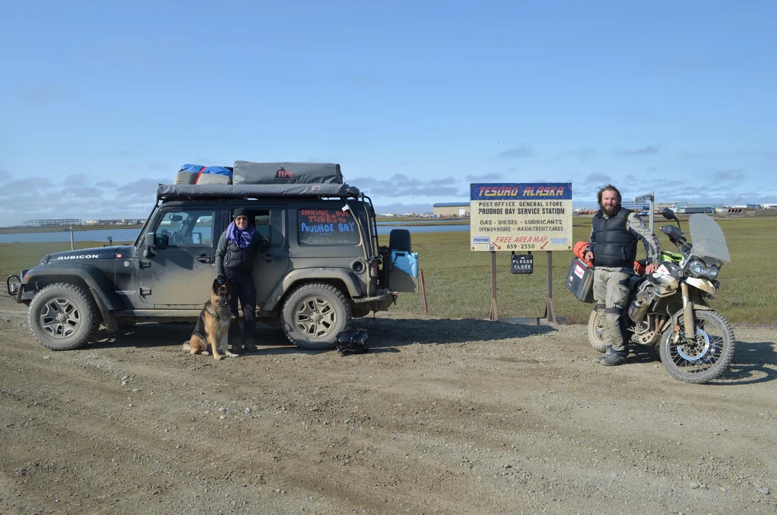 A Jeep with a rooftop tent and a loaded motorbike parked at the Prudhoe Bay service station sign in Alaska, with two riders and a German shepherd.