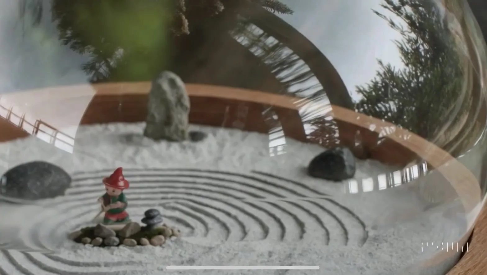 A miniature zen garden in a glass bowl, with raked white sand, small standing stones and a garden gnome figure holding a rake.