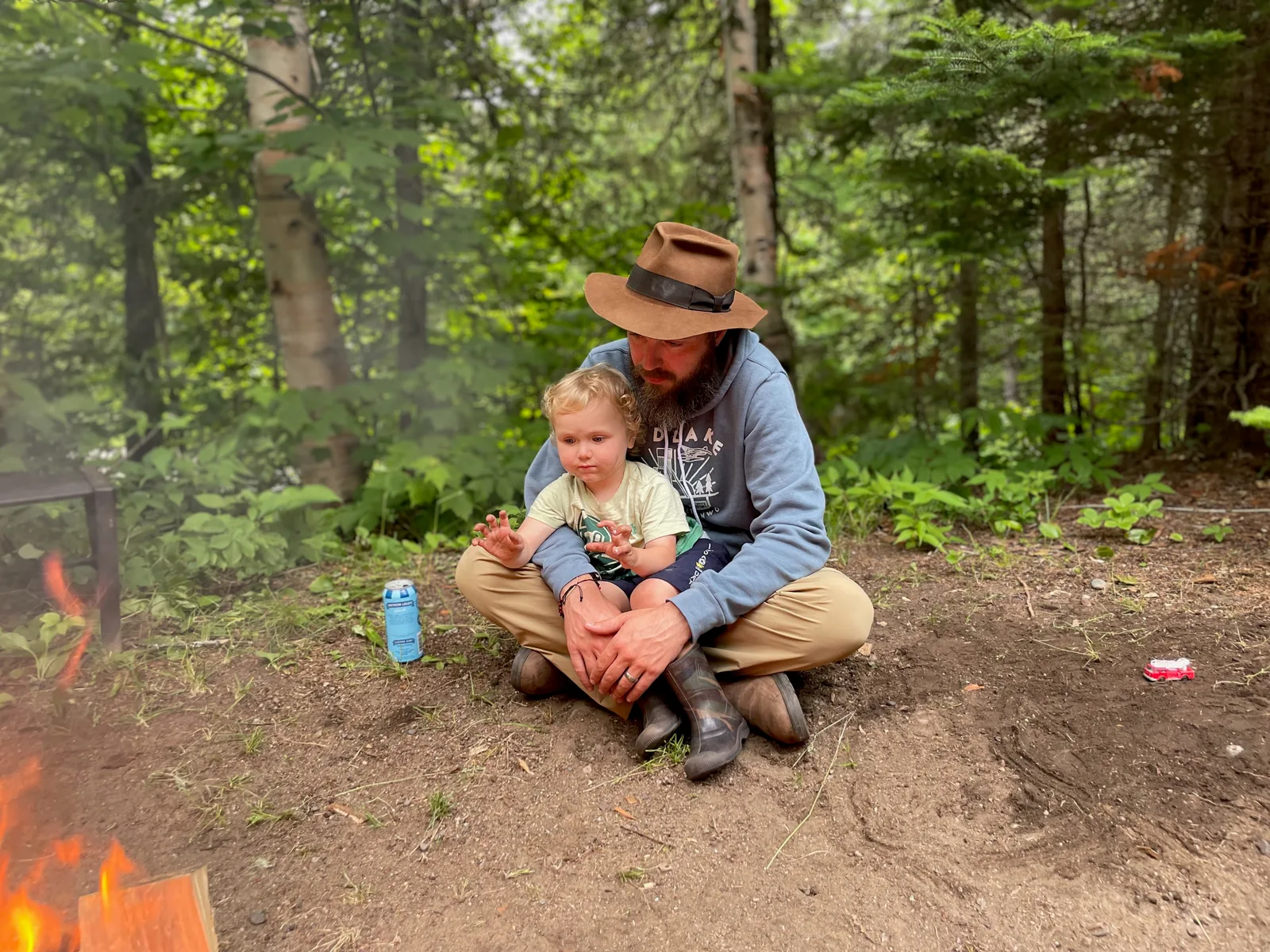 A man and a small child sitting next to a fire, the child is warming his hands by the fire.