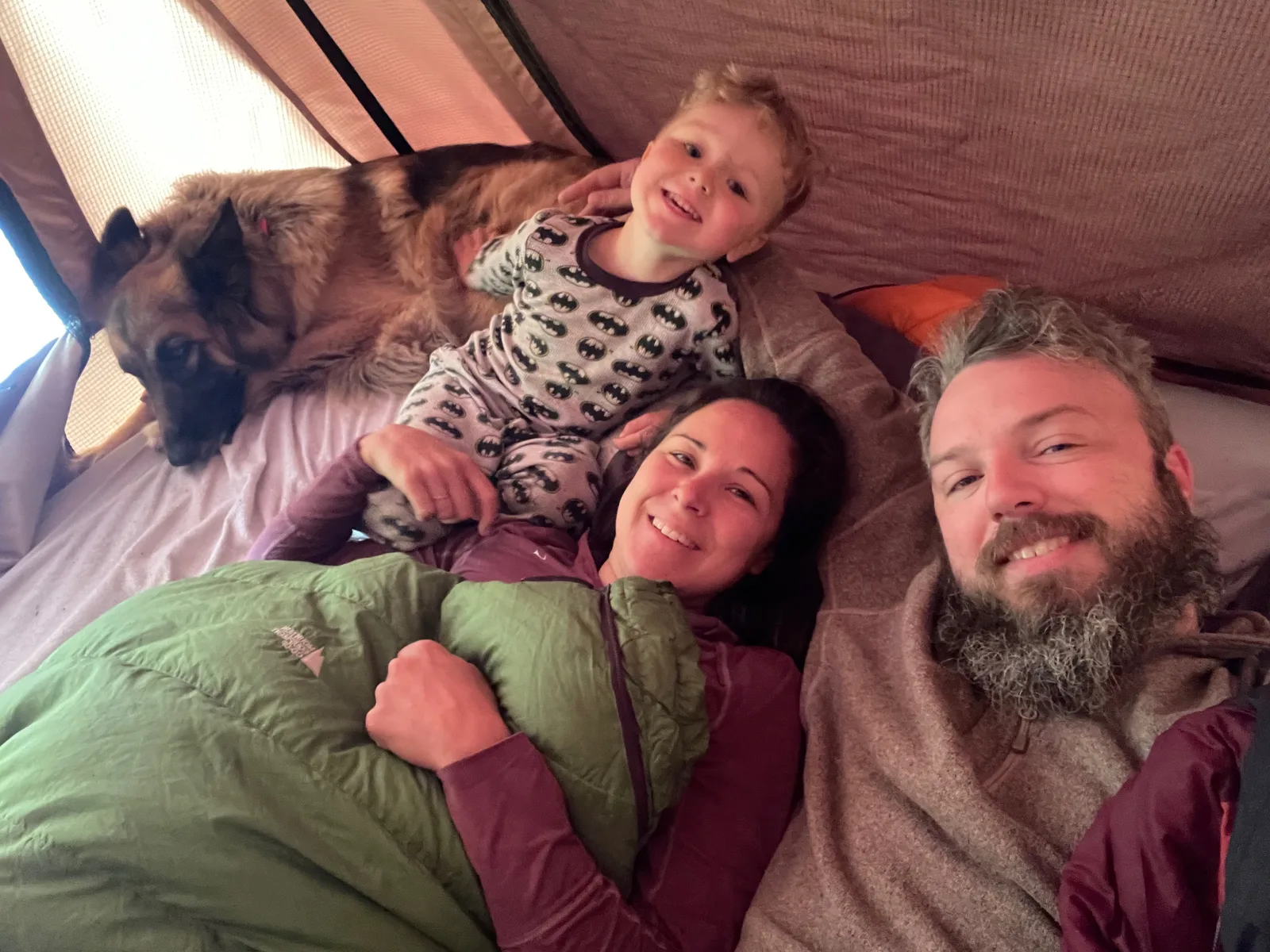 a family of three with a dog sleeping in a tent smiling at the camera.
