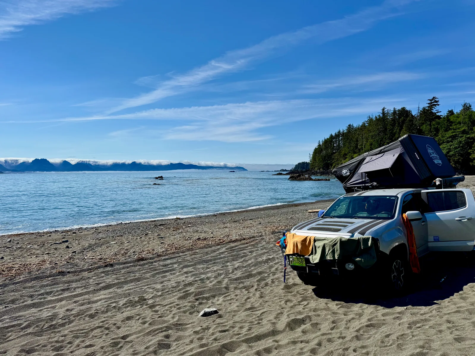 A pickup truck with a rooftop tent parked on a wild gravel beach, forest behind it and cloud-topped mountains across the water.