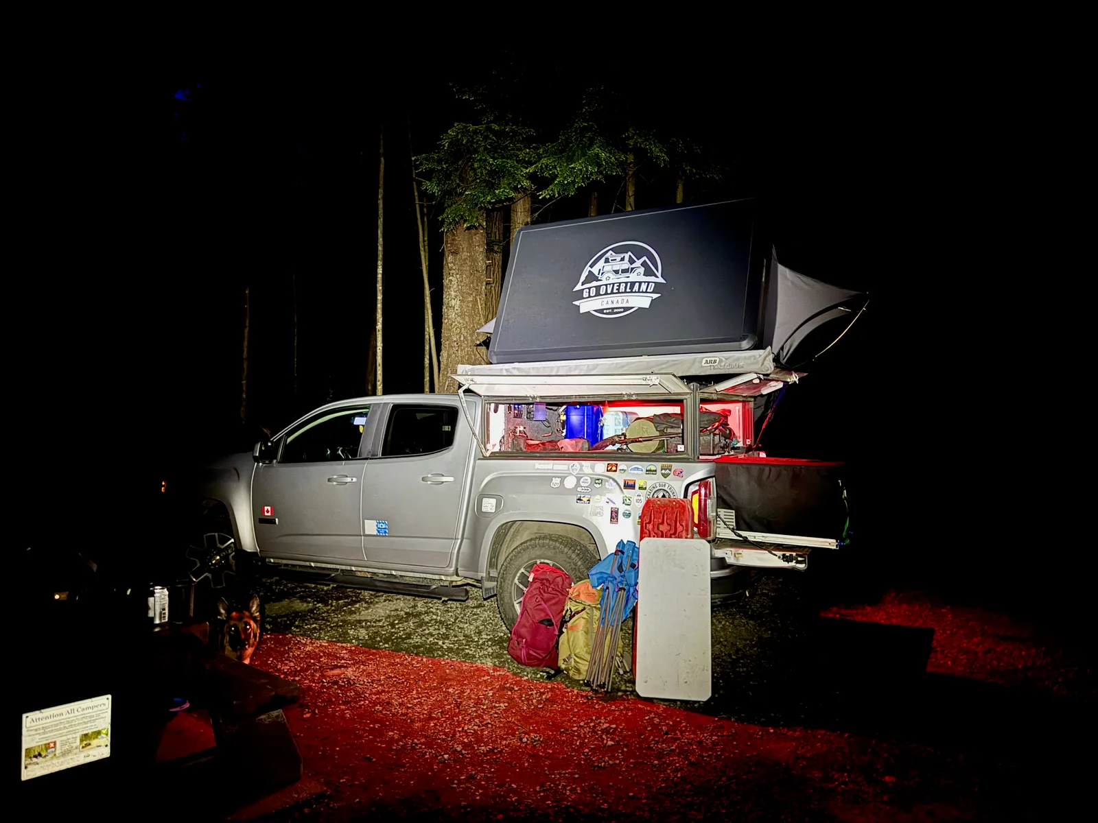 "Pac" the adventure truck, at night in a campground with a roof tent open and some backpacks leaned against the tire.
