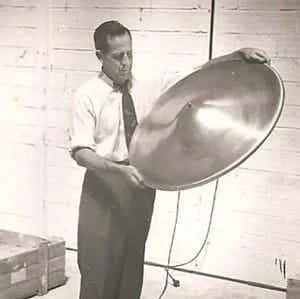 A vintage black-and-white photograph of a man in shirtsleeves holding up a large metal disc.