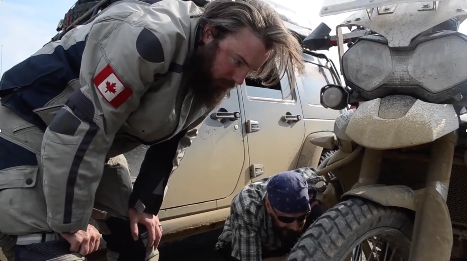 A motorcyclist staring at the front end of a muddy bike, with a muddy jeep in the background. Another man is crouched next to it wearing a bandana.