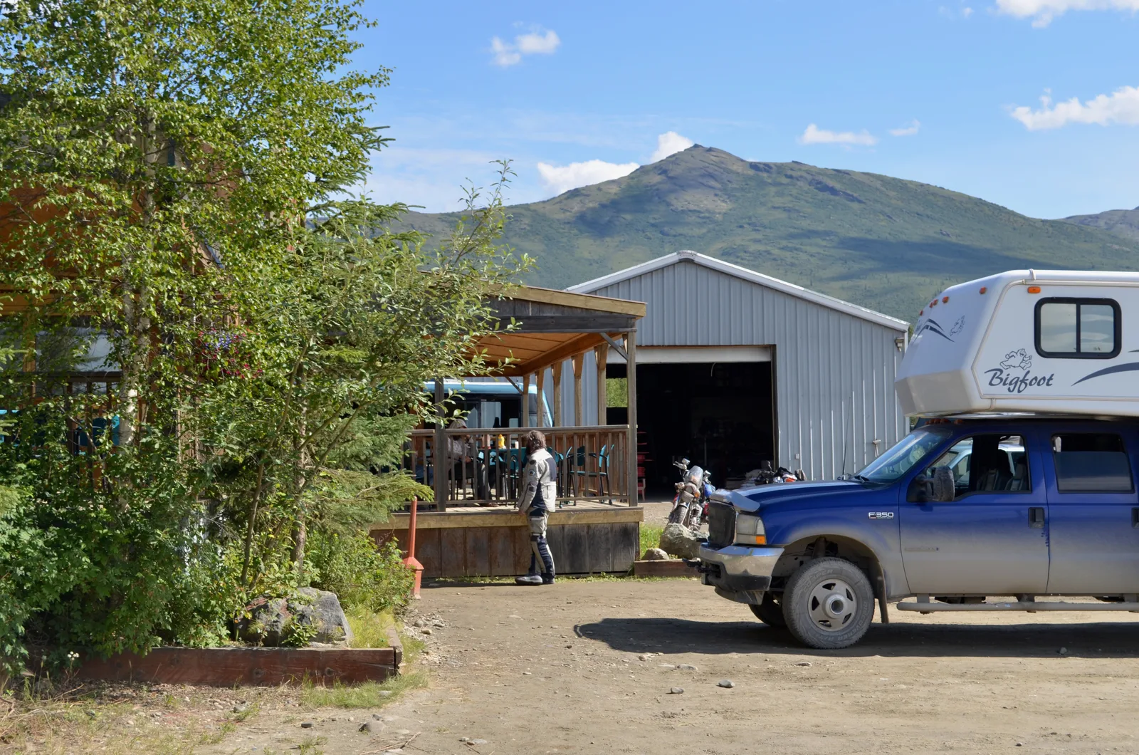 A wooden porch in front of a big mountain with a motorcyclist leaning against it.